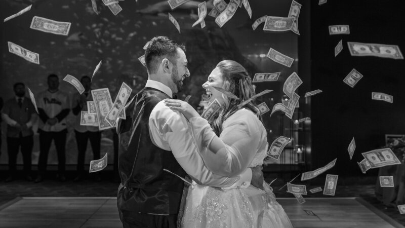 Bride and groom laughing together as they are showered with dollar bills during a Greek wedding tradition, capturing a lively celebration.