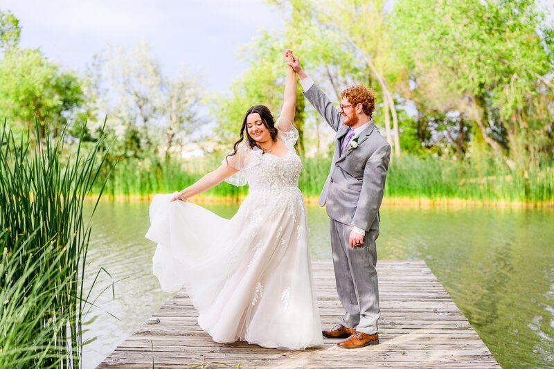 Bride twirling holding dress with groom holding her hand standing on dock with pond and green grass at The Windmill House in Chino Valley