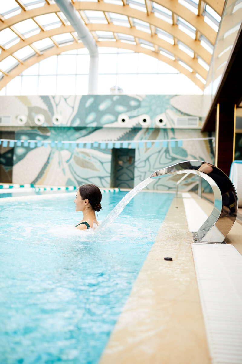 Person enjoying a pool with cascading water from a metal spout. The indoor pool area has a wooden arched ceiling and geometric wall patterns. Natural light streams in through large windows.