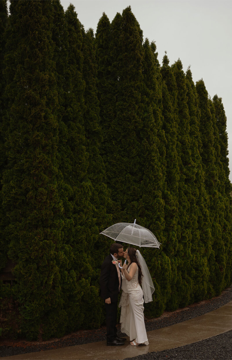 wedding couple kissing in the rain with umbrella in front of green trees