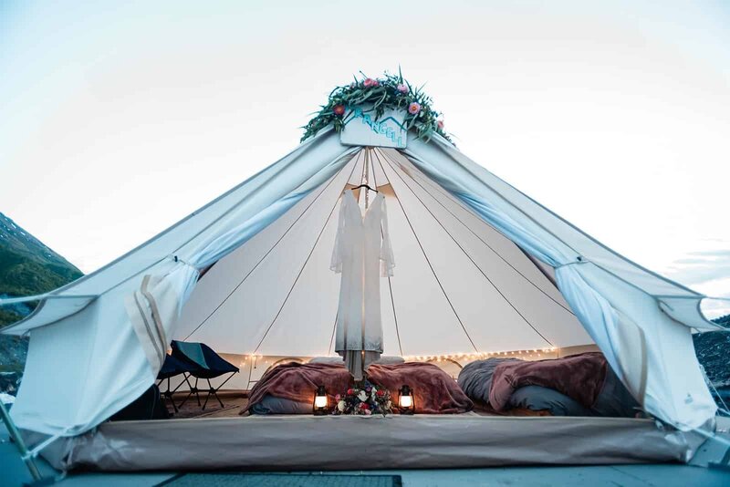 A light colored, luxury canvas bell tent is on a platform on the Matanuska Glacier in Alaska during a luxury glacier elopement .  There is a beautiful garland of colorful flowers on the tent door opening, and on the bouquet sits on the floor at the bottom of the wedding dress hem as it hangs displayed inside the tent, in front of the bed.  Small lanterns and twinkle lights in the tent add luxury touches to the elopement on the glaicer. 