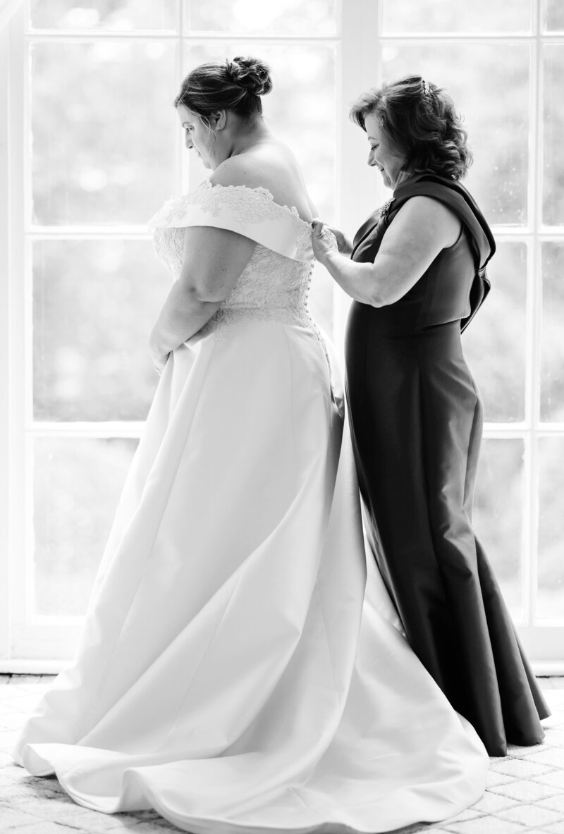 Mother zipping her daughter’s wedding dress in front of large windows, sharing a quiet, heartfelt moment before the ceremony.