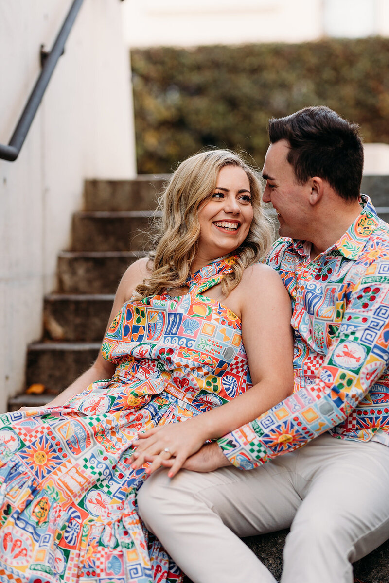 Bride to be looking and smiling at her husband to be while seated on stairs together in a New Acton laneway. Photography by Acacia & Grace.