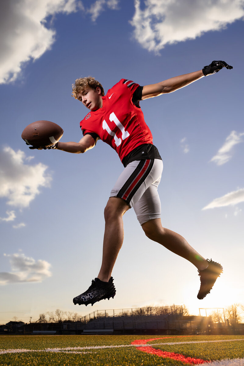 High school senior football player in a red jersey leaps midair, extending to catch a football at sunset on the field. Captured by a Dallas Senior Sports Photographer specializing in dynamic athletic portraits.