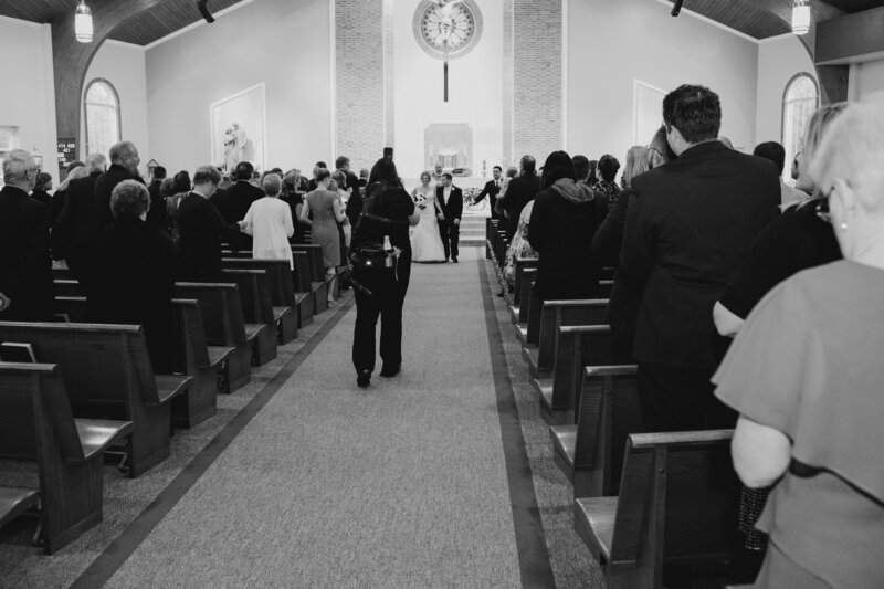 Black and white photo of Alexa Nahas photographing the bride and groom exiting the church, camera close to her face.