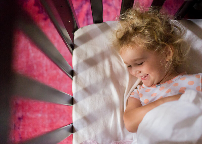 Curly haired toddler smiling in crib with pink rug in background