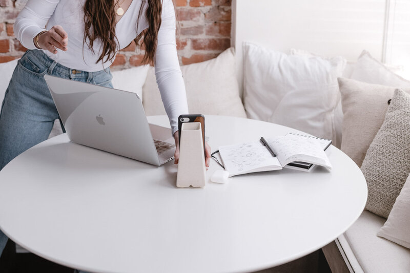 A girl in a white, long-sleeve crossbody top is placing a phone stand next to a Macbook pro laptop on a white table.