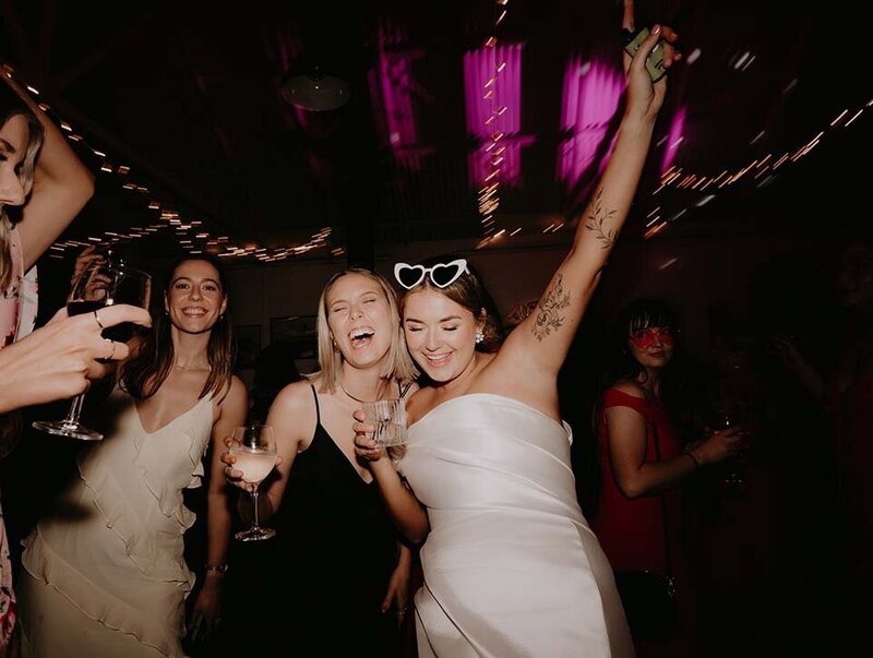 Bride in a white dress and heart-shaped sunglasses dancing with friends, holding drinks and surrounded by party lights.