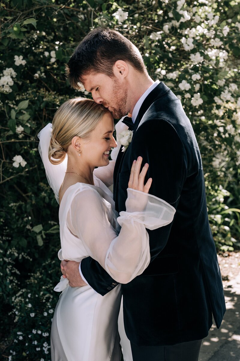 groom in tuxedo kisses bride in chiffon on forehead in front of white floral plants in garden at their christchurch cbd wedding