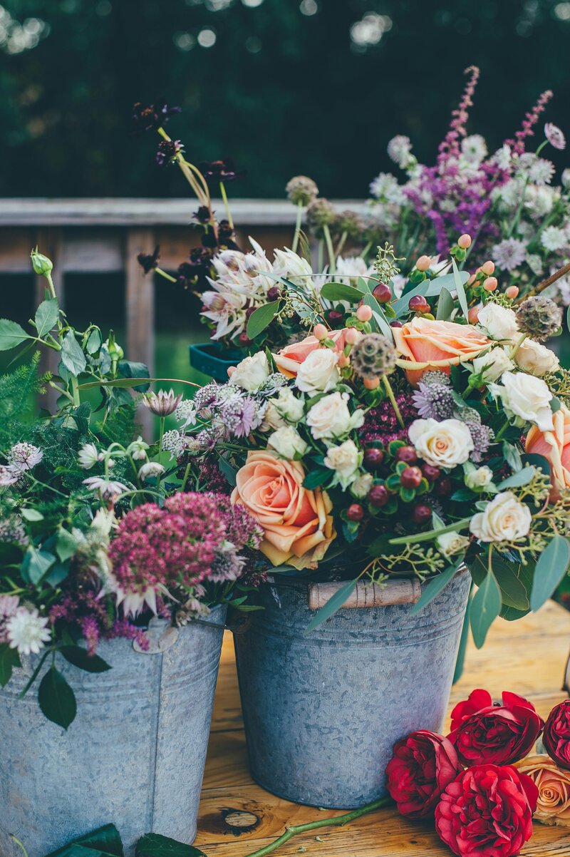 Assorted flower arrangements in rustic metal buckets, featuring roses, berries, and wildflowers in shades of peach, white, purple, and green, displayed on a wooden table outdoors.
