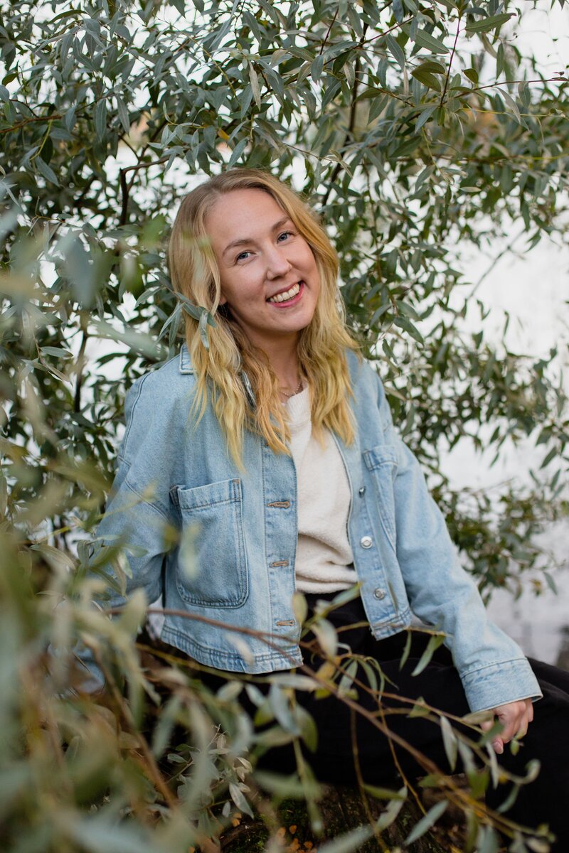 mielen paremminvoinnin ja kehomieliyhteyden valmentaja  istuu puunrungolla järvimaisema taustalla ja hymyilee