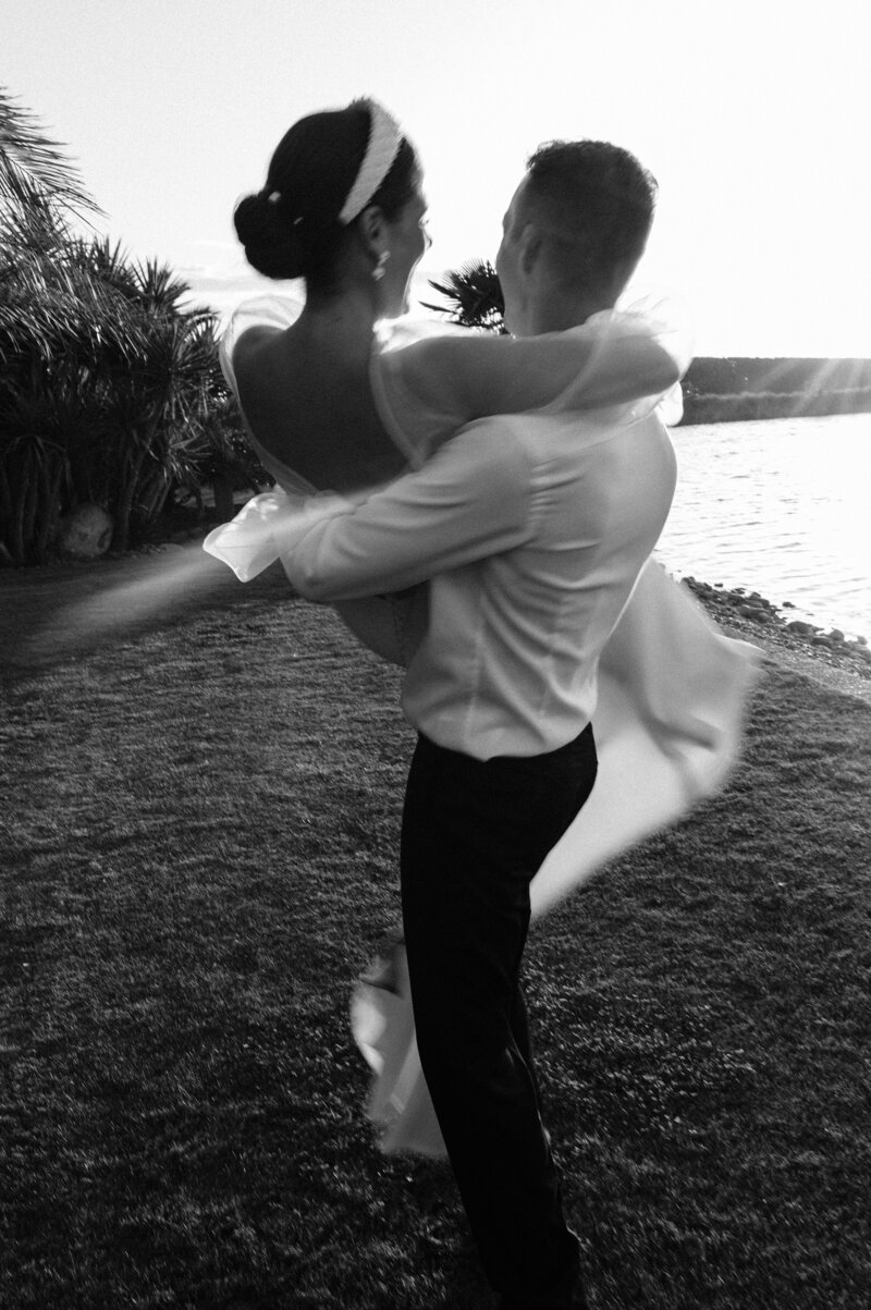 black and white shot of groom carrying bride blurry beside a lake at sunset in christchurch