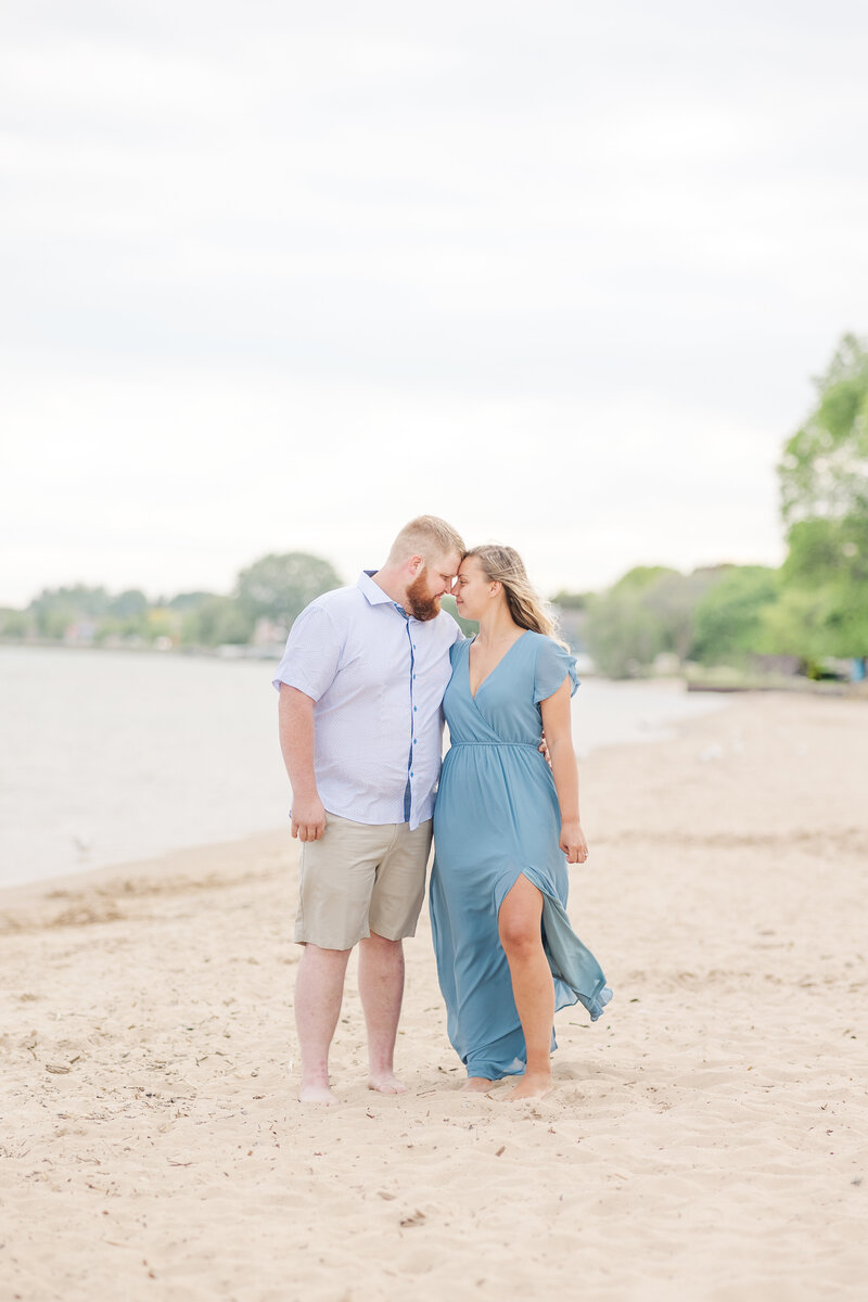 Newly engaged couple touches foreheads in a blue dress and button down shirt while standing on a windy beach