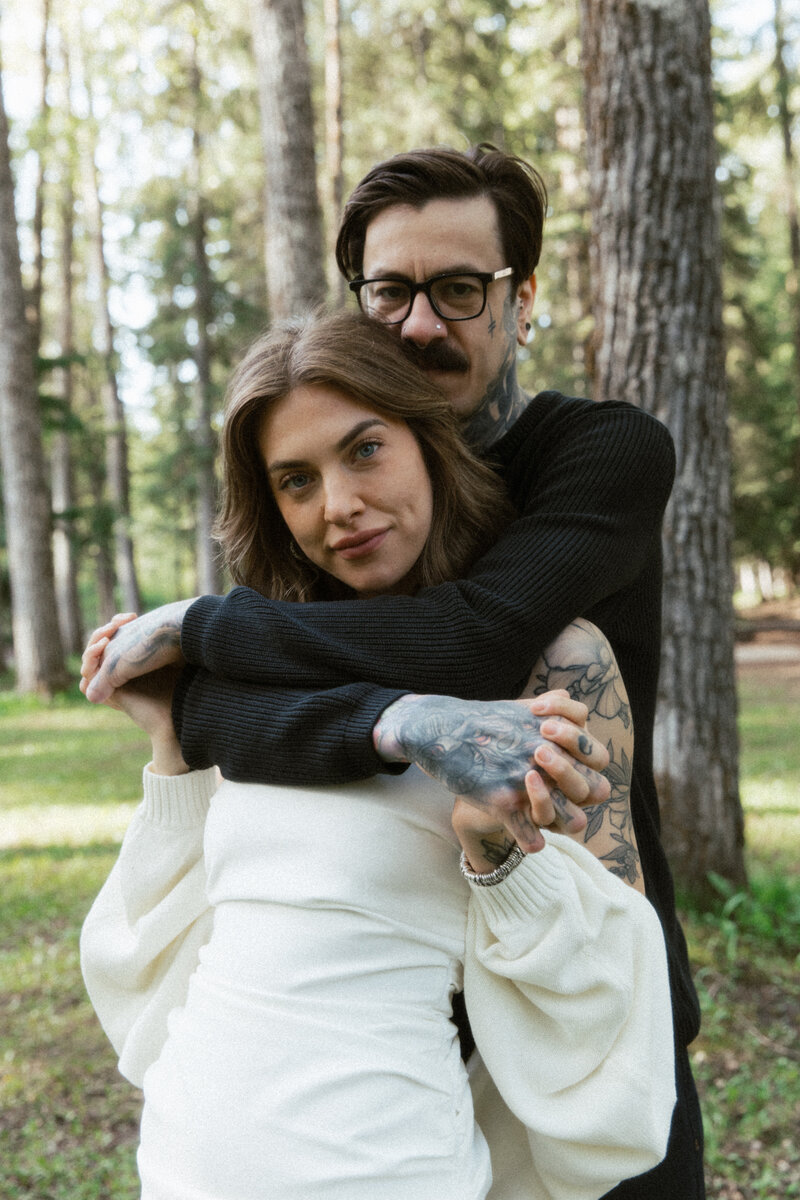 Young couple with tattoos snuggle up with each other looking directly at the camera during Alberta engagement session