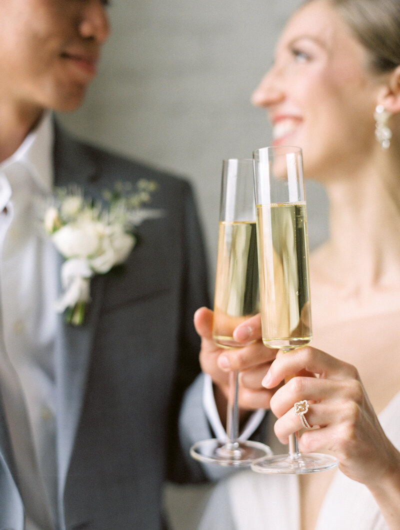 a bride and groom in the background looking at each other with champagne glasses and their hands toasting to one another in the foreground