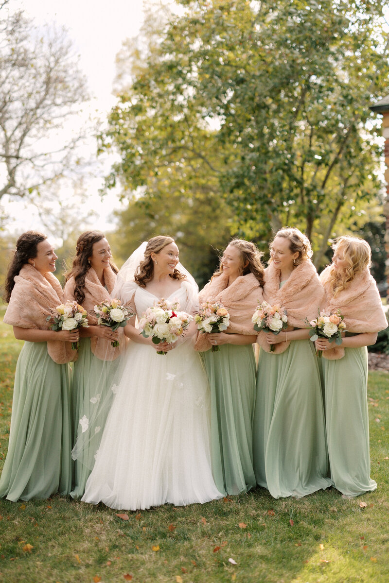 November wedding at Graylyn Estate, designed and planned by Charming Carolina — bride and bridesmaids in sage green gowns and fur shawls posing for a group photo outside the historic estate.