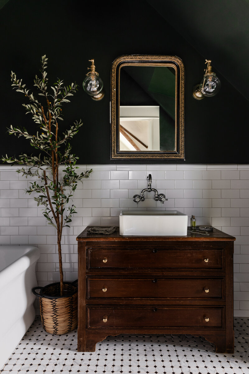 A European inspired guest bathroom with vintage vanity, dark green walls, and classic subway tile. Black and white marble floors.
