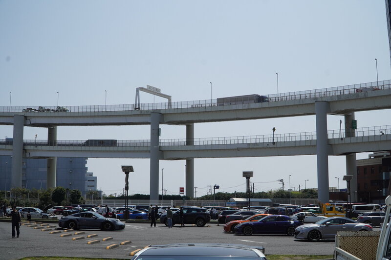 Wide view of Daikoku Parking Area during the day, showcasing rows of sports and luxury cars with elevated expressways above — a prime meetup spot for Tokyo car enthusiasts.