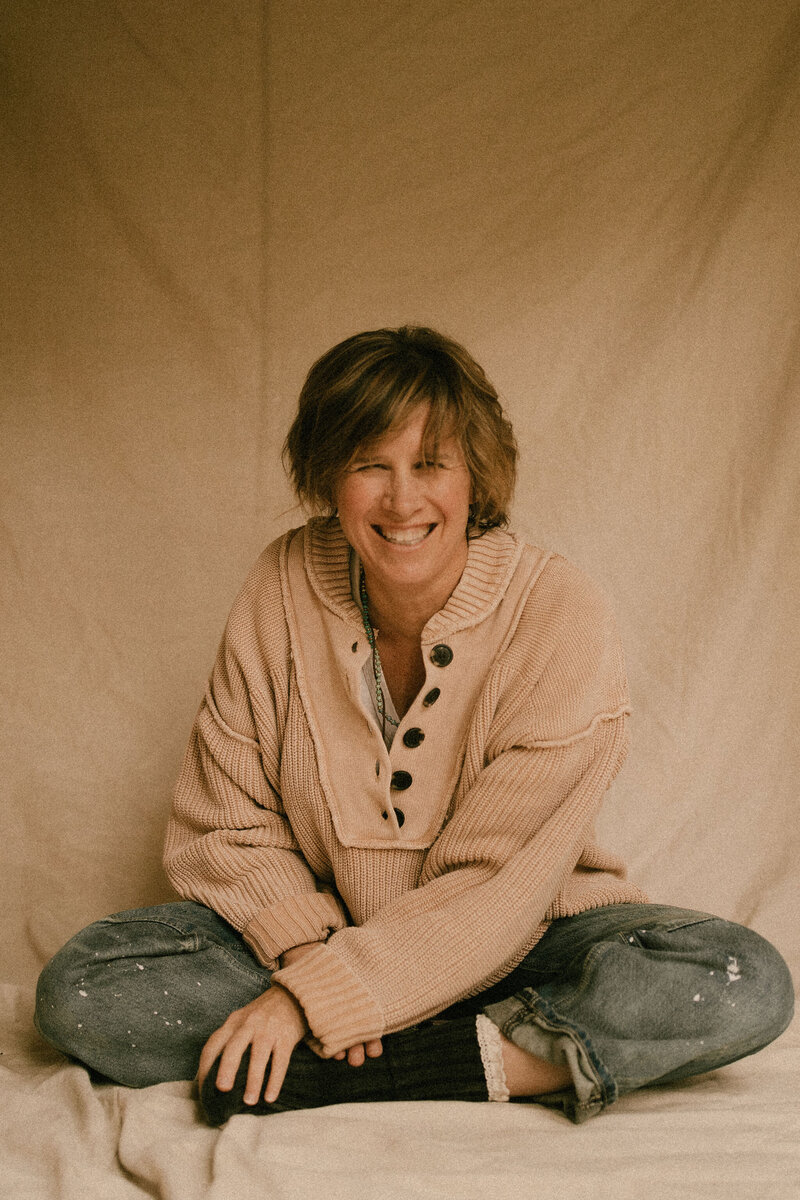 Photographer Melanie Dalton sitting in front of a canvas background wearing jeans and a sweater. 