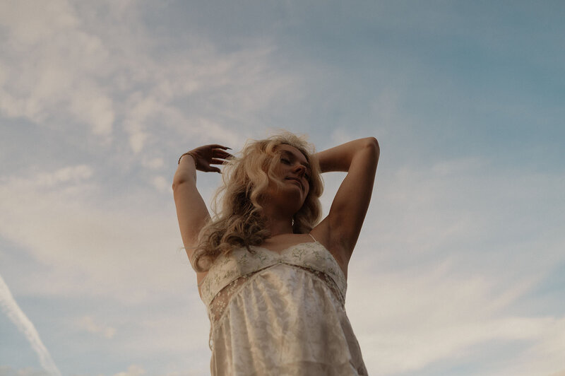 woman posing for a photograph in the outdoors against the blue sky with clouds wearing a flowy top