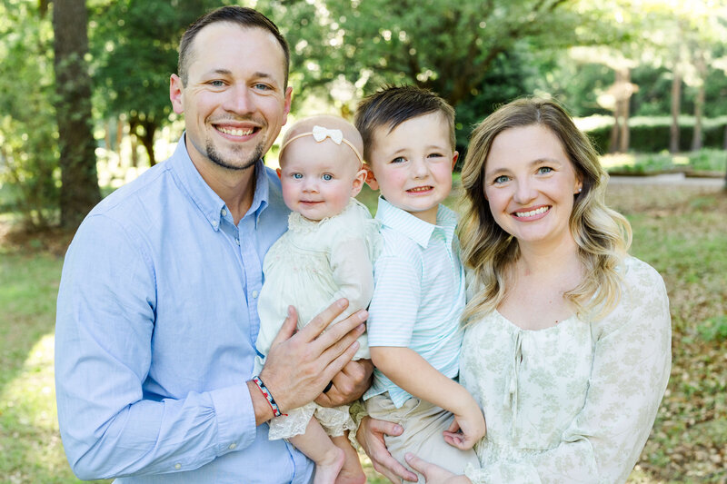 Family of 4 standing closely together while parents hold the siblings and smile at the camera in dothan alabama park