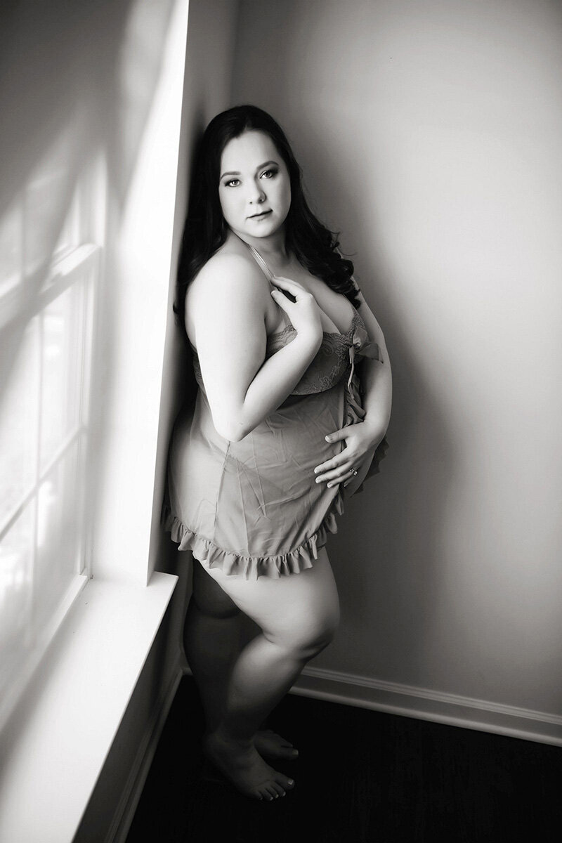 A woman in lingerie stands by a window, softly lit in black and white. She gazes confidently at the camera, exuding a sense of empowerment and grace.