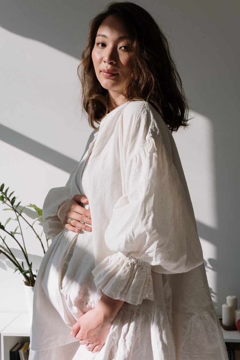 Expecting mother frames her pregnant belly with her hands while the sun  shines in through the window