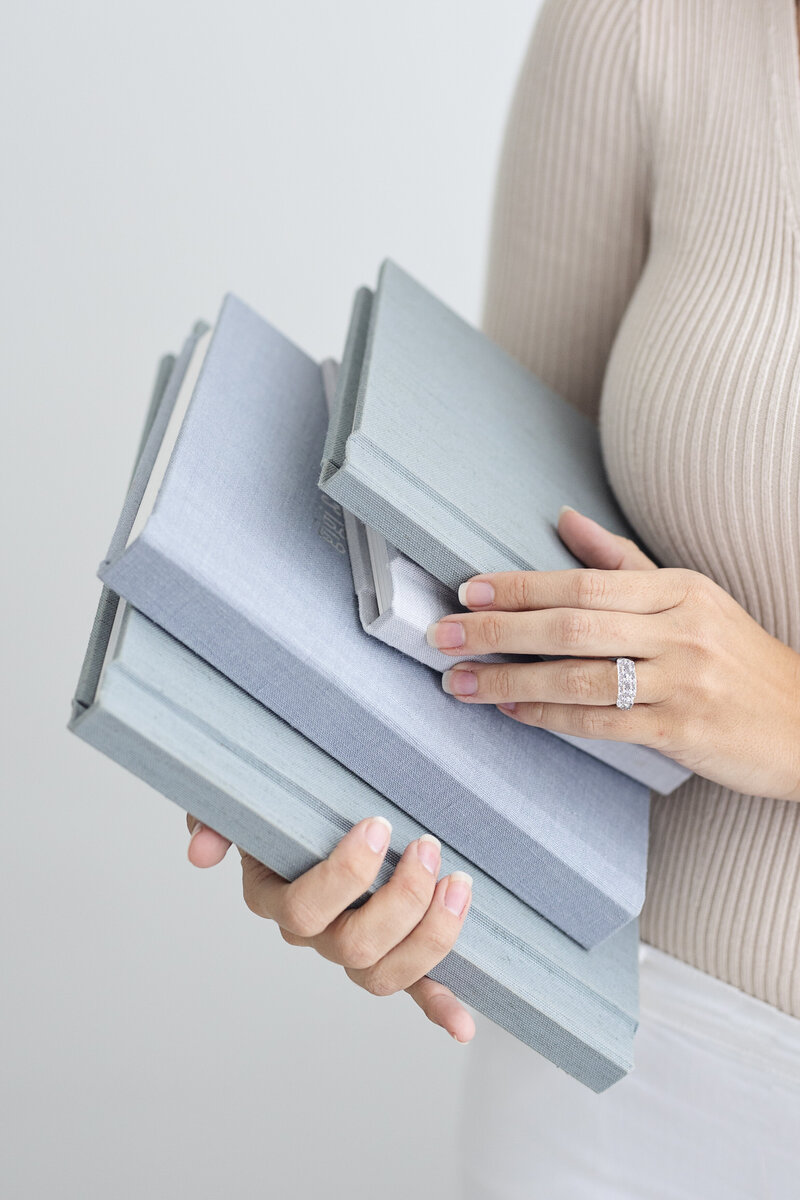 A close-up of a woman's hands holding a stack of photo albums that are different shades of blue holding them against her side wearing a beige sweater.