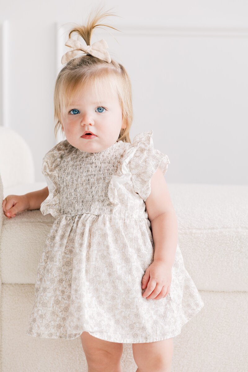 little girl stands holding onto couch in a neutral dress during her photography session in charlotte nc