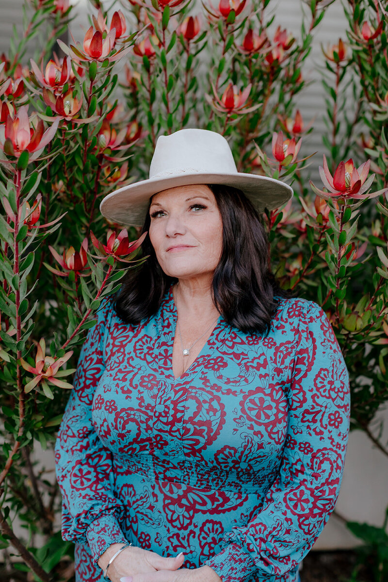 Close-up outdoor portrait of Susie Schumacher wearing a wide-brimmed hat and a blue patterned dress, standing in front of tall red and green flowering plants with a calm, confident expression – Susie Schumacher Life Coach