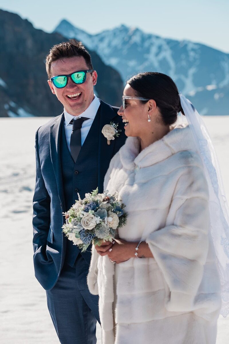 A bride and groom in their wedding finest laugh and smile after just having exchanged elopement vows on the glacier during their two day adventure luxe Alaska elopement.   The sky is bright blue and the snow gleams white against the dark mountains rising up from the glacier.  The bride holds her bouquet and the groom is looking to the camera and smiling a huge smile, his eyes covered by mirrored blue sunglasses that match his navy blue suit. 