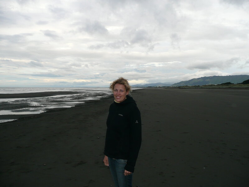 Therapeutin mit gewelltem blondem Haar schwarzer Fleecejacke und Jeans am Strand in Neuseeland mit Bergen im Hintergrund bedecktem Himmel und Wellen