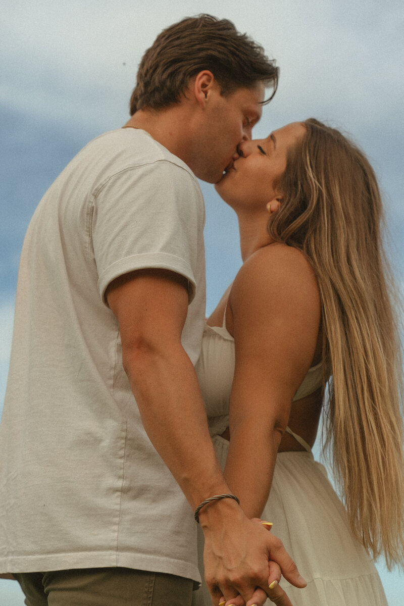 Close-up of a couple sharing a tender kiss while holding hands, set against a soft blue sky during their outdoor photo session.