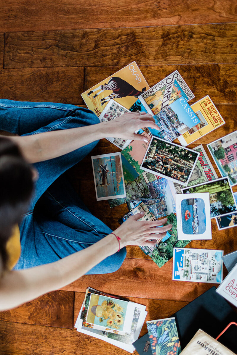 Person sitting on a wooden floor, surrounded by various postcards, some depicting travel destinations