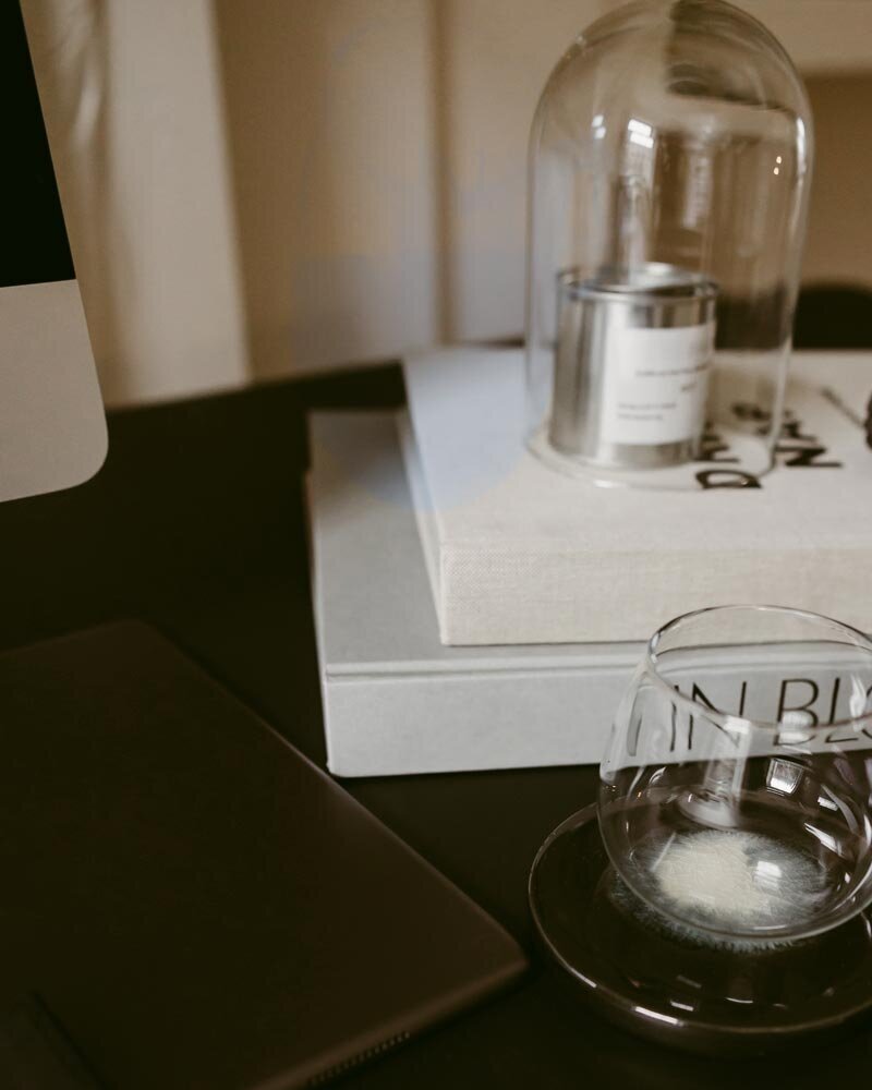 A modern desk setup with stacked books, a candle under a glass dome, a tablet, and an empty glass on a saucer. The atmosphere is minimalist and cozy.