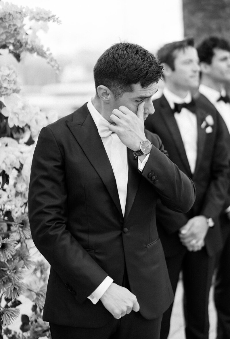 Groom wiping away a tear in a black and white candid photo during the ceremony, captured in documentary wedding style