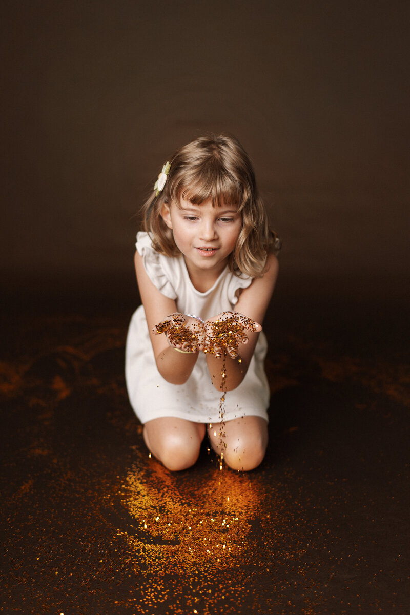 A girl kneels in front of a black background while glitter pours from her hands. 