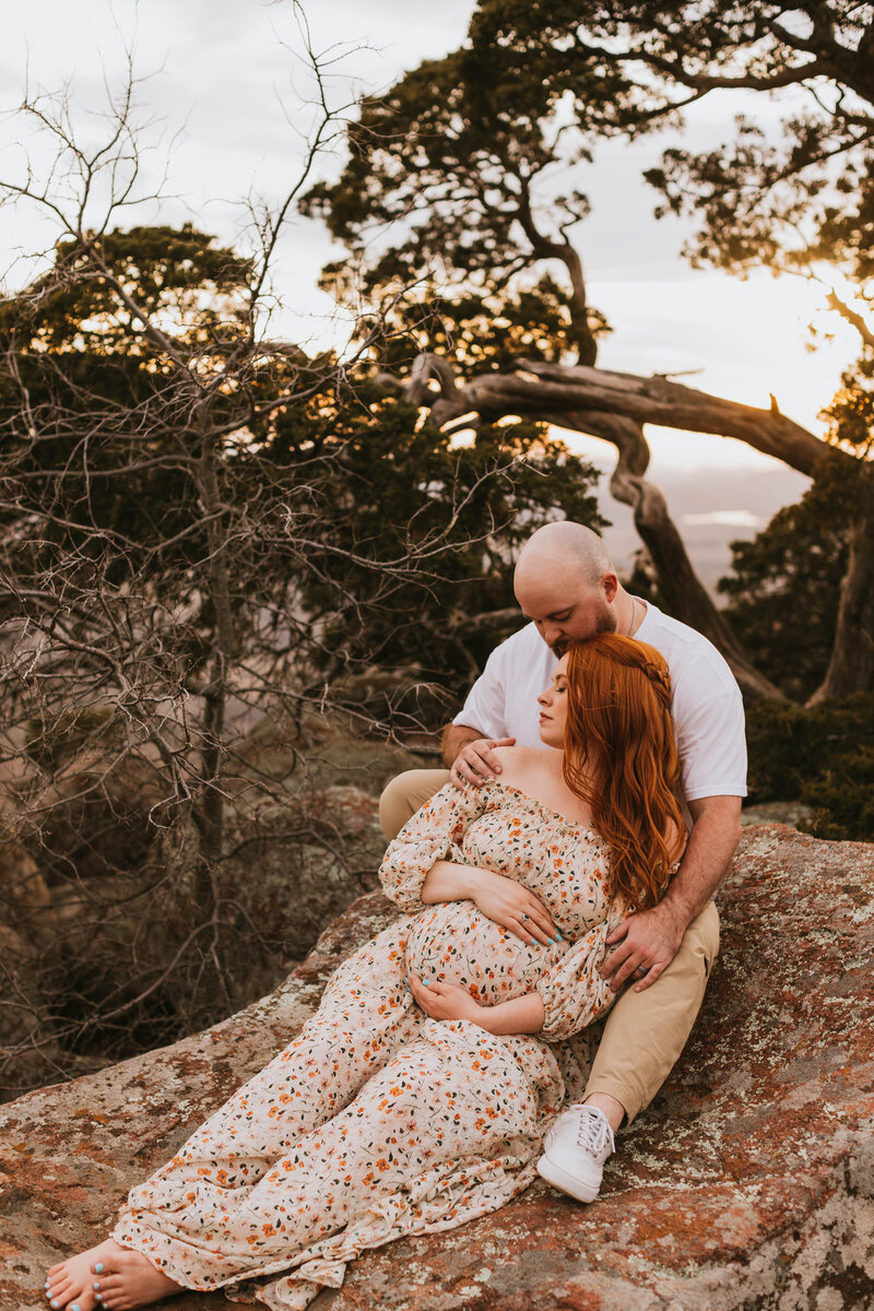 Expecting couple sitting on a large rock and mom is wearing a floral print dress