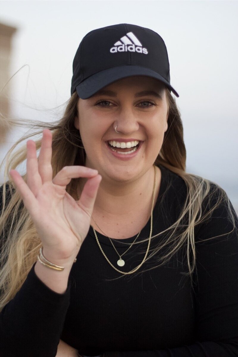 Michaela Jade in a black top & baseball cap smiling showing the okay sign 