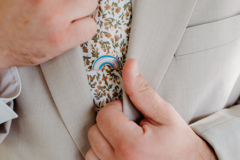 A person adjusting their jacket with a focus on a trans pride pin on their tie.