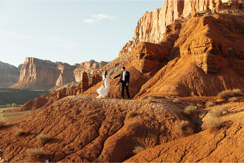 A distant shot of a bride and groom holding hands and looking at each other. They are standing on red rock with stunning red rock and buttes behind them for hundreds of miles.