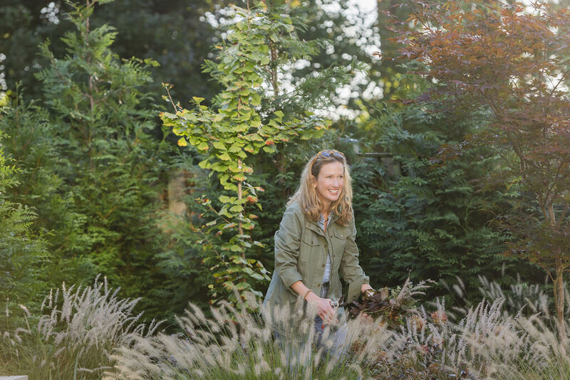 Woman with light brown blonde hair wearing army green jacket pruning grass in private backyard