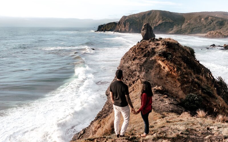 Couple holding hands on dramatic New Zealand cliffside overlooking the ocean during engagement photoshoot by Zanthe Vorsatz.