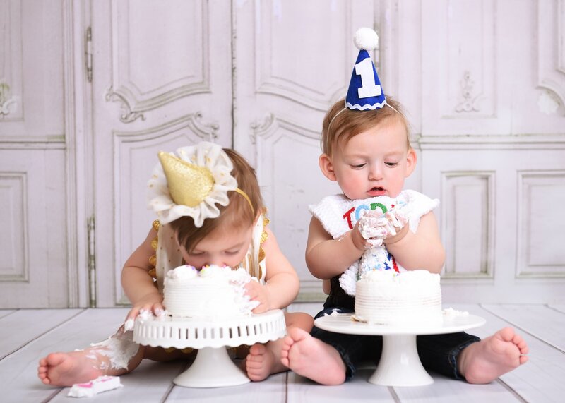 Twin babies celebrating their first birthday with a cake smash; one wearing a gold party hat diving into the cake and the other in a blue "1" hat curiously examining the frosting, set against a vintage white background.