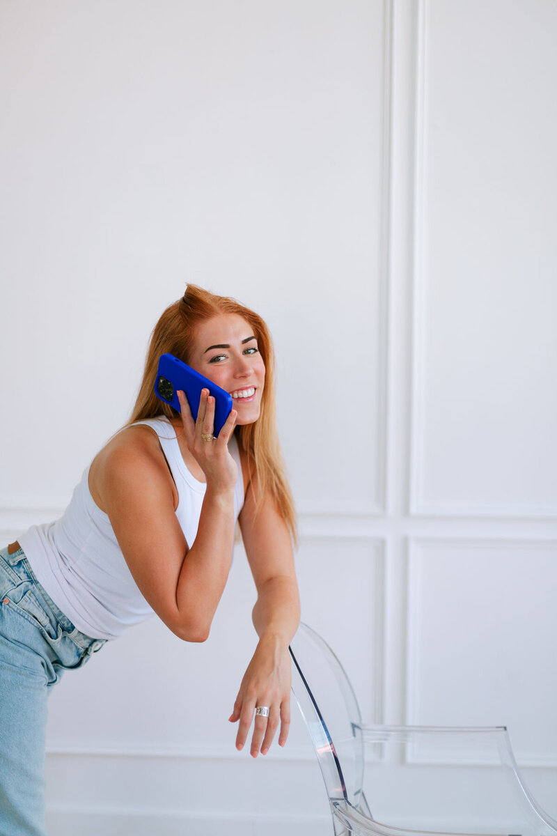 ALT: Grace sitting on floor with arms crossed over knee holding phone smiling at camera with papers on floor under her and taped to wall behind her