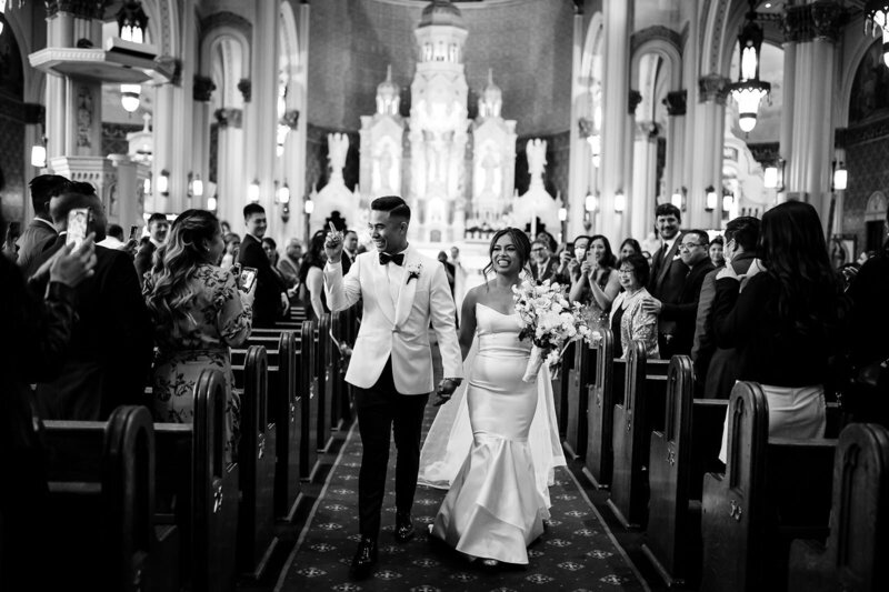 Professional San Francisco Bay Area wedding photography documenting authentic ceremony recessional as bride in elegant mermaid dress and groom in white dinner jacket walk down aisle of Saints Peter & Paul Church in North Beach with applauding guests, ornate altar, and Gothic architecture captured in classic black and white