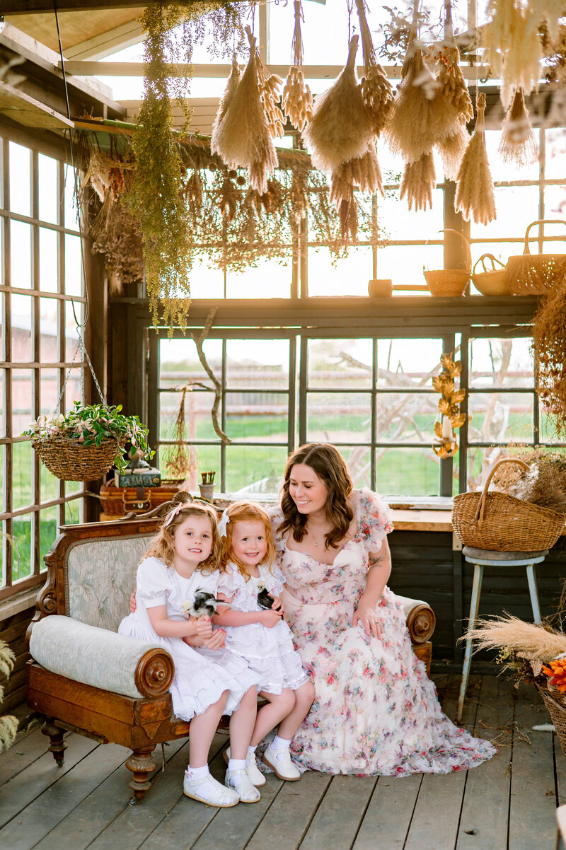 Mother and two young daughters sitting together in a rustic sunroom surrounded by dried florals and natural light, photographed by Jennifer L. Kirk Photography in Allen, Texas.