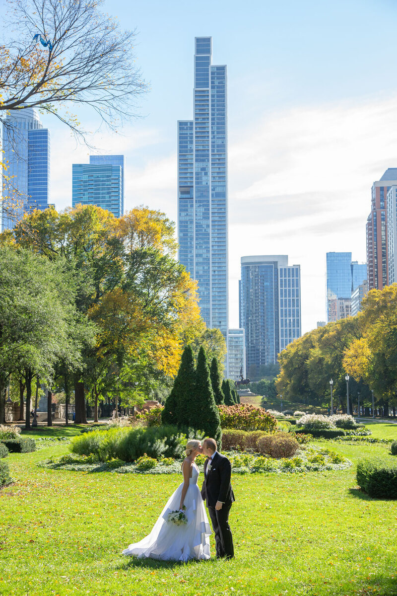 Bride and groom kissing in an outdoor sitting with greenery and tall buildings behind