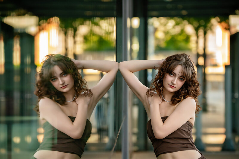 A high school senior girl poses in front of a mirrored building so that it shows her reflection.