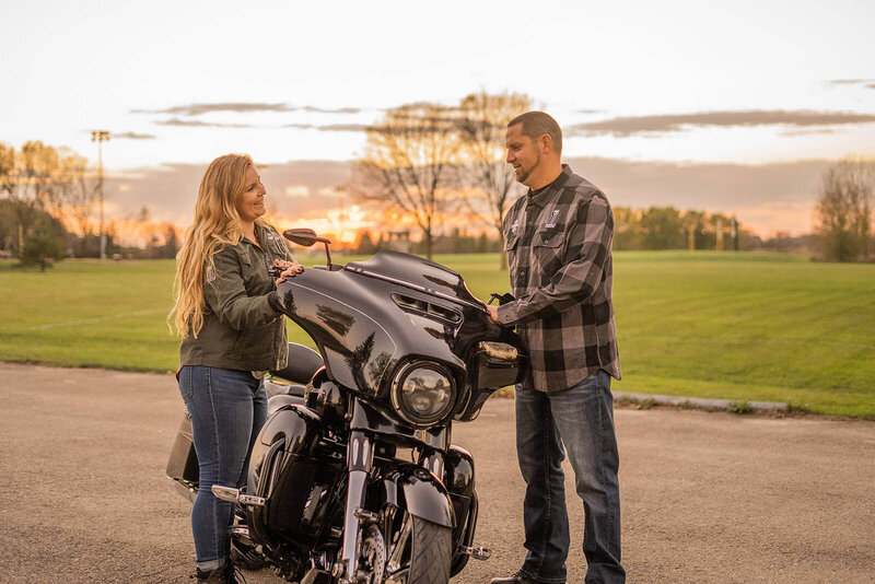 husband and wife laughing while standing by harley davidson motorcycle at sunset